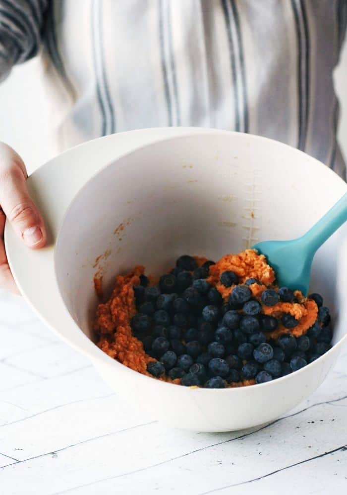 hands mixing a bowl of sweet potato muffin batter with blueberries