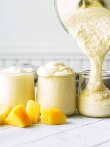 3 cups being filled with a creamy yellow pineapple smoothie surrounded by chunks of pineapple