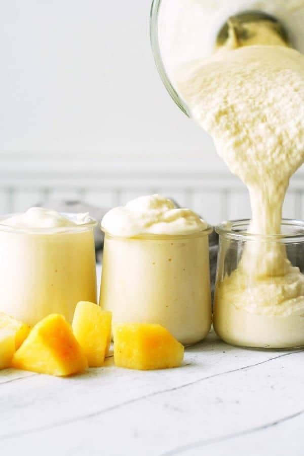 3 cups being filled with a creamy yellow pineapple smoothie surrounded by chunks of pineapple