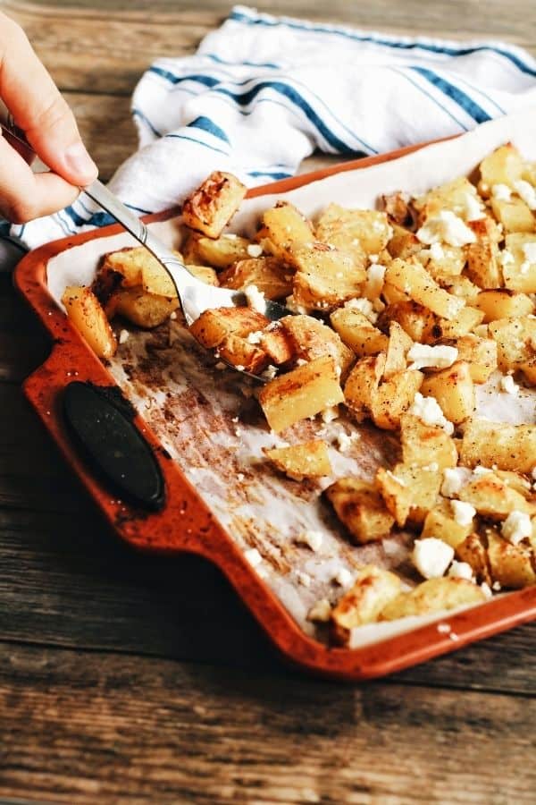 hand scooping up some garlicky roast potatoes with feta of a baking sheet