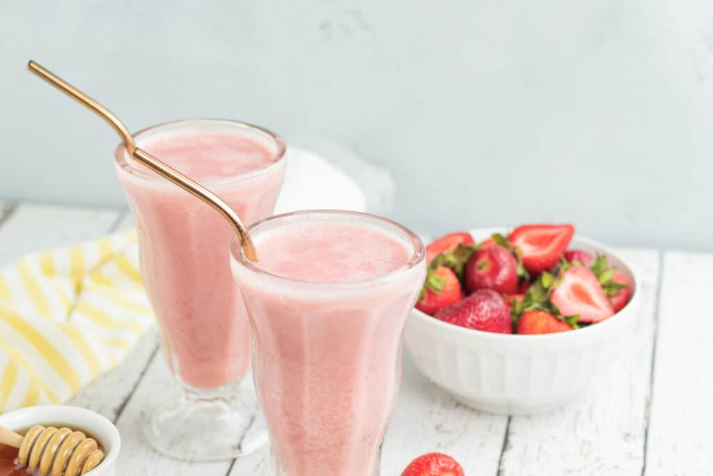 two strawberry smoothies in glasses with gold straws with a white and yellow towel and a bowl of honey to the left and a bowl of strawberries to the right 