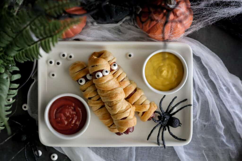 crescent roll mummy hotdogs on a white plate with ketchup and mustard on either side with spiders 